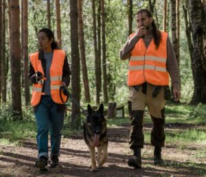 Two people with rescue dog wearing safety vests in forest.